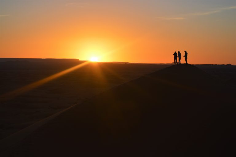 3 tourists stand on top of a sand dune as the sun rises behind them